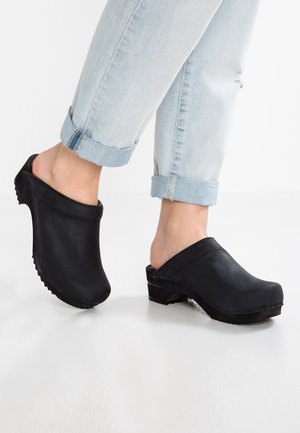 Mustat nahkaiset puukengät, joissa on pyöreä kärki, avoin takaosa ja matala korko, jossa teksturoidut pohjat. Vasileveysiset vaaleat farkut.