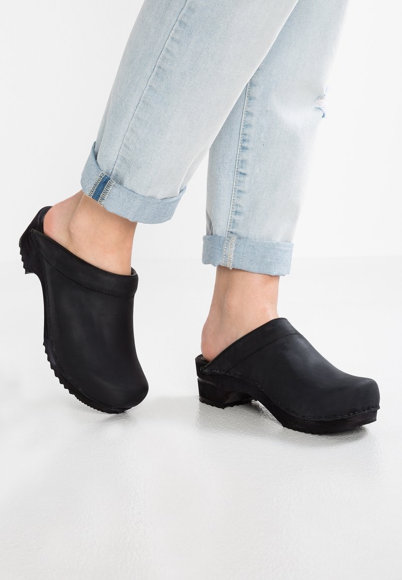Sabots noirs en cuir lisse, dotés d'une pointe arrondie, d'un dos ouvert et d'un talon bas avec des semelles texturées. Jean en denim clair longueur mollet.