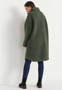 Vrouw in een lange groene fleece jas, blauwe jeans en zwarte laarzen, staande zijwaarts tegen een witte muur.