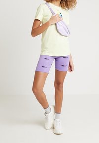 Kvinna klädd i en ljusgul T-shirt, lila Nike Air cykelshorts, vita sneakers, grå strumpor och en matchande lila axelväska.