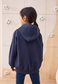 Temno modra oversize kapuca s kapuco, rebrastimi manšeti in robom. Mehak material, vidni šivi, kombinirana s temno modrimi hlačami ob svetli steni.
