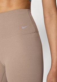 Visoke tajice, priljubljene tijelu, u prigušenoj bež boji s glatkom teksturom. Sadrže mali sivi Nike logo na pojasu.