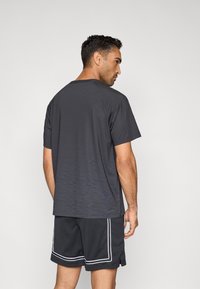 Σκούρο γκρι αθλητικό μπλουζάκι με διαβαθμισμένο σχέδιο και κοντά μανίκια, συνδυασμένο με μαύρο σορτς από διχτυωτό ύφασμα με λευκές ρίγες στα πλαϊνά.