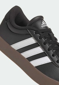 Czarny syntetyczny sneaker z białymi paskami, brązową gumową podeszwą, teksturowaną cholewką i czarnymi sznurowadłami. Zawiera logo Adidas na języku.