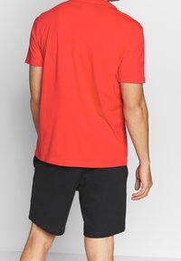 Rotes Baumwoll-T-Shirt mit Rundhalsausschnitt und kurzen Ärmeln, kombiniert mit schwarzen Shorts. Der Stoff wirkt glatt und leicht.