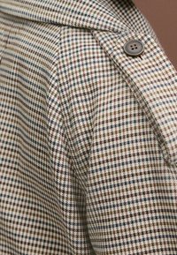 Geruite stof in beige, bruin en blauwe tinten met een gladde textuur; heeft een schouderknopdetail en een klassiek kraagontwerp.