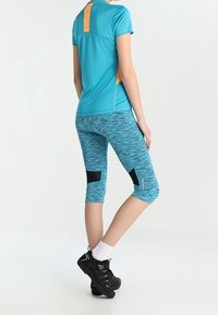 Tyrkysové atletické tričko s oranžovými akcenty, spárované s modrými vzorovanými legínami po kotníky s černými panely, doplněné černou obuví.