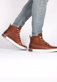 Zapatillas altas de cuero marrón con suela de goma blanca, punta redondeada, cordones beige y acabado texturizado, que presentan un logotipo en el lateral.