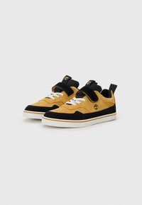 Chaussures en daim jaune et noir, dotées d'une semelle blanche plate, de lacets contrastants et d'une sangle Velcro pour un ajustement sécurisé. Logo Timberland présent.