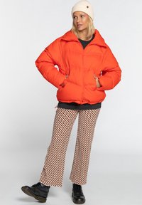 Giacca puffer arancione brillante con colletto alto, due tasche frontali e una vestibilità ampia, abbinata a pantaloni svasati marroni e bianchi a fantasia.