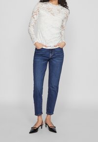 Top bianco in pizzo a maniche lunghe con motivi floreali e scollatura a onda, abbinato a jeans in denim blu e décolleté nere a punta.