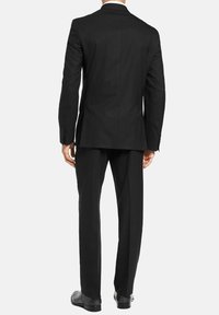 Veste de costume noire avec une fente unique, design ajusté et manches longues. Les pantalons présentent une coupe droite. Les deux sont fabriqués en tissu lisse.