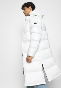 Cappotto piumino lungo e bianco con cappuccio, dotato di sezioni trapuntate, chiusura con cerniera frontale e uno spacco laterale per facilitare i movimenti.