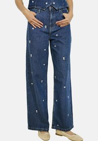 Džinsa plati kāju bikses ar zilu mazgājumu, izkaisītu baltu ziedu izšuvumu un augstu vidukli ar kabatām. Gluda tekstūra, ikdienišķs dizains.