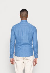 Camisa de manga larga azul claro de tela de lino con cuello puntiagudo, puños abotonados y dobladillo curvado. La vista trasera muestra una textura suave.