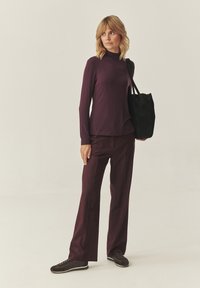 Langsleeve, fitted paarse top gecombineerd met losse, bijpassende paarse broek. Zwarte tote bag, donkere sneakers en een neutrale achtergrond.