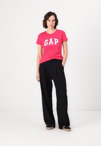 Pink bomulds T-shirt med hvid "GAP" logo, parret med høj talje sorte plisserede bukser og sorte sneakers, stående på en ensfarvet baggrund.