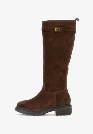 Vysoká hnědá semišová bota s nízkým klínovým podpatkem, podrážkou s drážkami a ozdobným popruhem se zlatou přezkou blízko vrchu.