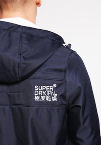 Giacca con cappuccio blu navy in tessuto liscio, con logo ricamato bianco sul retro. Include un cordino e cuciture visibili.