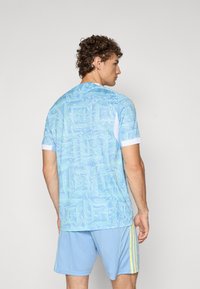 Lyseblå treningstrøye med et virvlende mønster, korte ermer og hvit krage, kombinert med lyseblå shorts og gule detaljer.