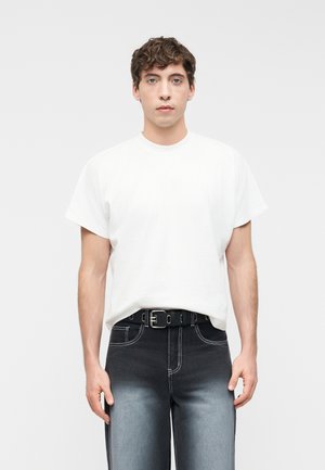 Giovane uomo che indossa una maglietta bianca semplice infilata in jeans neri sbiaditi con cuciture bianche e una cintura nera, in piedi contro uno sfondo neutro.