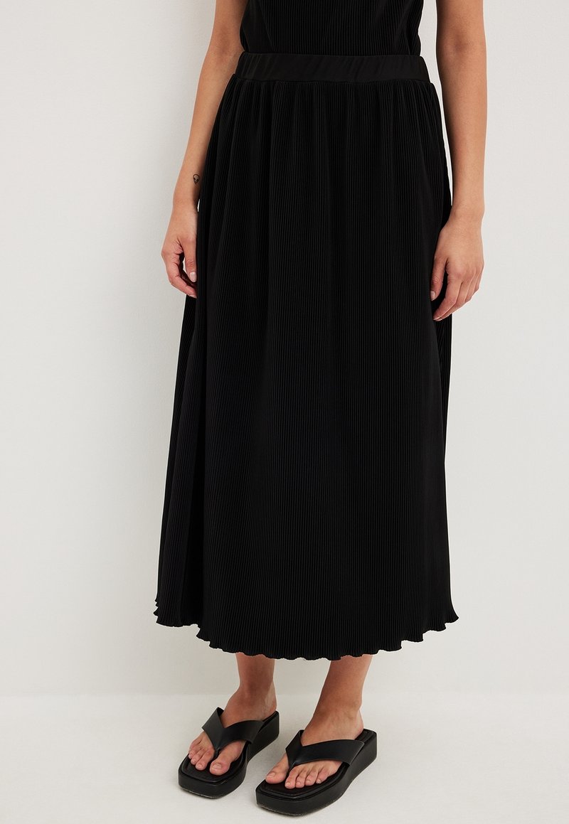 Femme portant une jupe midi plissée noire et des sandales à plateforme noires, debout devant un fond clair uni.