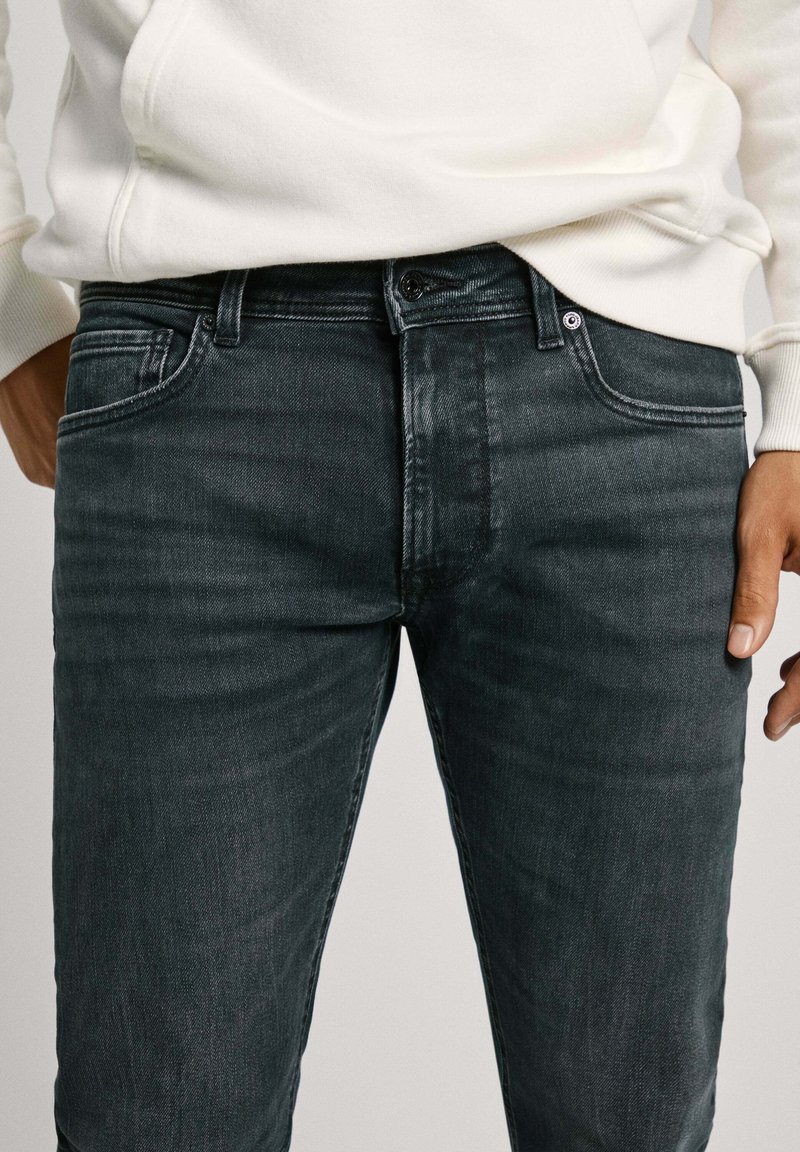 Dunkelgraue Denim-Jeans mit geradem Schnitt, fünf Taschen, dezenten Waschungen und einem Knopfverschluss. Tragen mit einem weißen Hoodie.