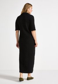 Robe maxi noire texturée à manches courtes, avec une coupe ample et une silhouette droite, associée à des chaussures vert olive.