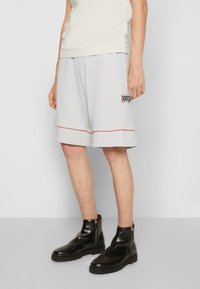Pantaloni corti grigi chiari con una linea di accento rossa, dotati di una toppa logo nera sul lato. Abbinati a stivaletti neri con cerniera alla caviglia.
