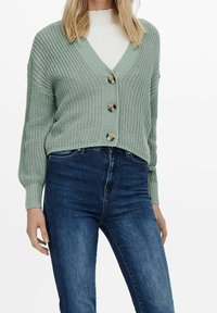 Cárdigan de punto verde menta con escote en V, cinco botones grandes y puños acanalados, combinado con jeans de mezclilla azules. Tejido texturizado y corte relajado.