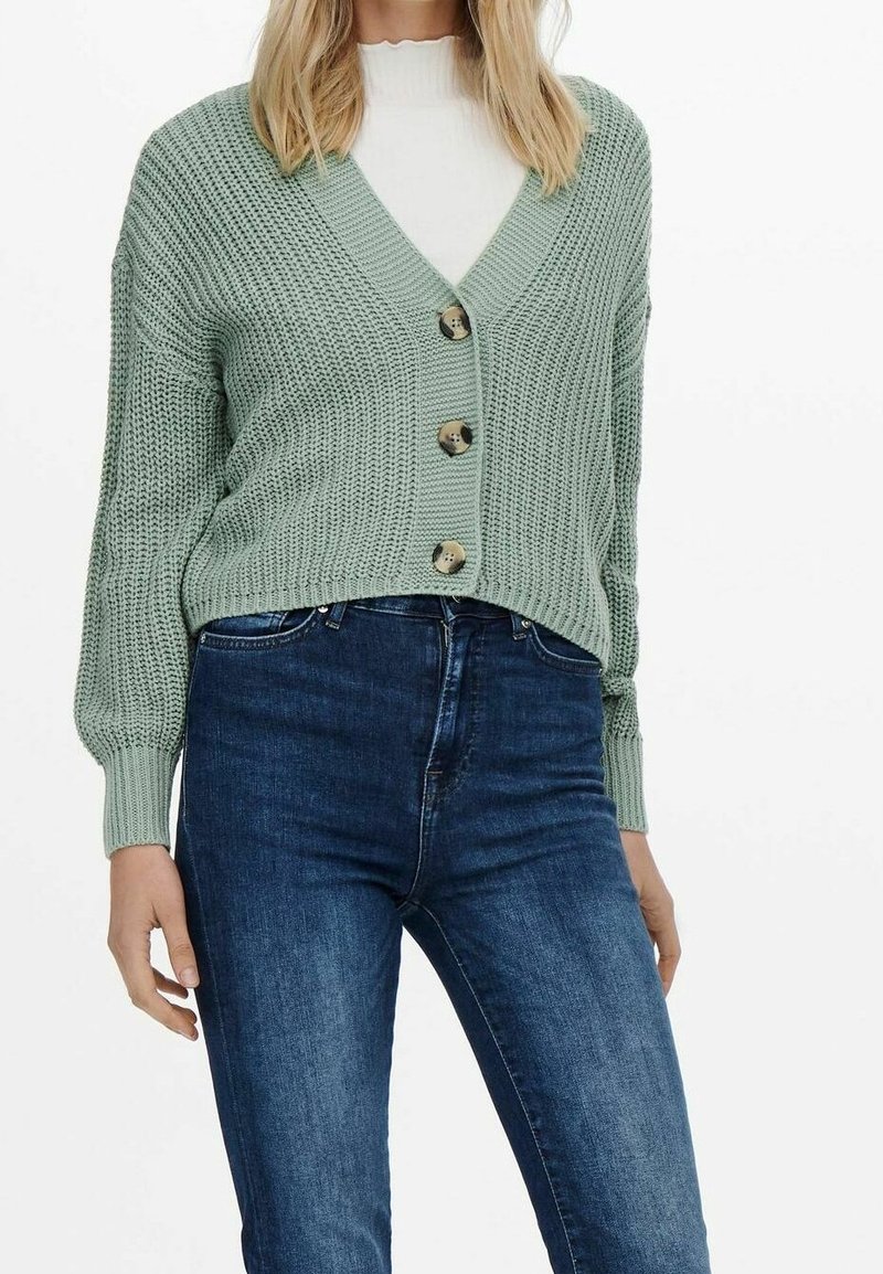 Cárdigan de punto verde menta con escote en V, cinco botones grandes y puños acanalados, combinado con jeans de mezclilla azules. Tejido texturizado y corte relajado.
