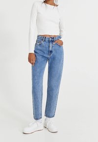 Osoba v modrých džínech s vysokým pasem, bílém dlouhém cropped topu, bílých ponožkách a bílých teniskách, stojící na pozadí bez vzoru.