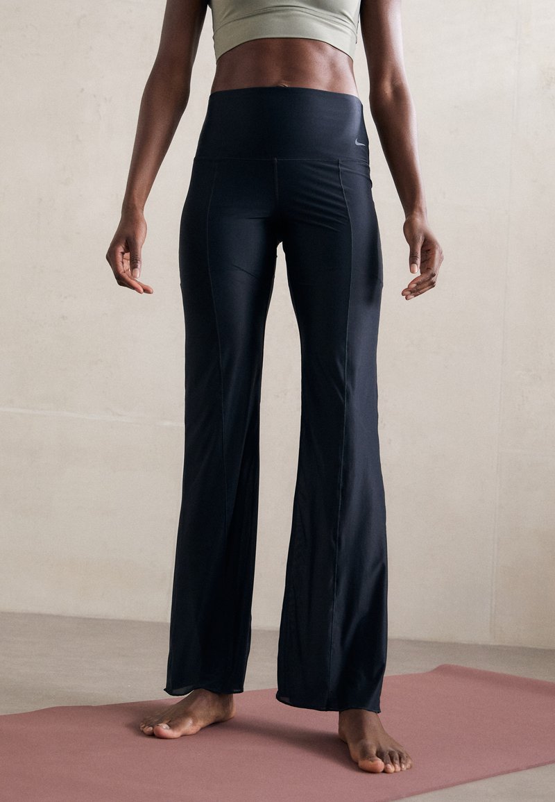 Sieviete stāv basām kājām uz jogas paklāja, ģērbusies melnās flared leggingos un gaišas krāsas sporta krūtītes, pret vienkāršu sienu.