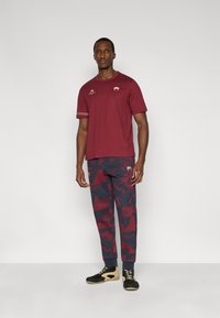 Μπορντό κοντομάνικη μπλούζα με χρυσές λεπτομέρειες, συνδυασμένη με σκούρο μπλε παντελόνι με κόκκινο αφηρημένο σχέδιο. Μαύρα και χρυσά υποδήματα ολοκληρώνουν την εμφάνιση.