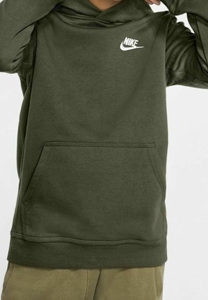 Personne portant un sweat à capuche Nike vert olive avec une poche frontale, le logo visible sur la partie supérieure gauche de la poitrine, les mains ajustant la capuche.