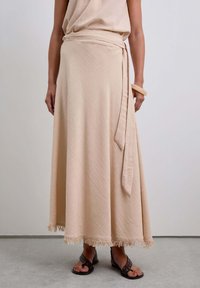 Beige Wickelrock aus strukturierter Stoff, mit einem Bindeband in der Taille und Fransen am Saum. Trägt dazu braune Sandalen mit Nieten.