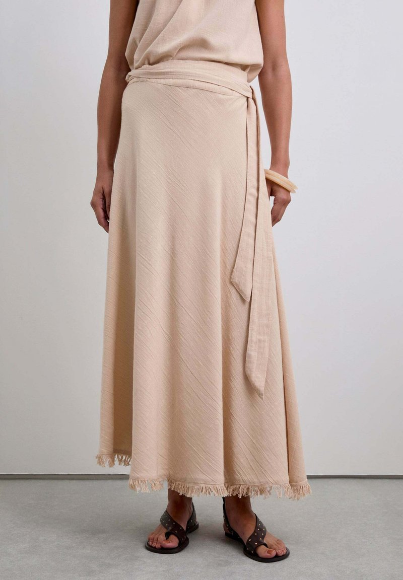 Beige Wickelrock aus strukturierter Stoff, mit einem Bindeband in der Taille und Fransen am Saum. Trägt dazu braune Sandalen mit Nieten.