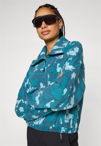 Teal-Fleece-Pullover mit halbem Reißverschluss, abstraktem Muster und hohem Kragen. Mit Branding auf der Brust und dem Ärmel sowie einer strukturierten Oberfläche.