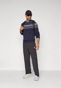 Tumši zils džemperis ar baltām un zilām rakstām, melnas bikses, pelēkas sporta kurpes, melna mugursoma un cepure. Izskatu papildina saulesbrilles.