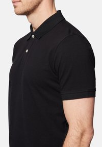 Czarna koszulka polo z teksturowanej tkaniny, z rypsowym kołnierzykiem i krótkimi rękawami. Posiada dwa jasne guziki na listwie.
