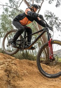 Horský bicykl s černým rámem a červenými vidlicemi, vybavený silnými pneumatikami se vzorem dezénu, jede přes skok z hlíny v lesnaté oblasti.