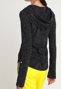 Kvinna iklädd en svart melerad huva långärmad topp i kombination med ljusgula byxor, visad bakifrån och från sidan.