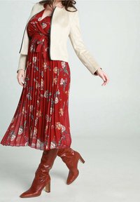 Veste en daim beige sur une robe plissée à fleurs rouges, longueur genou. Bottes en cuir marron à talons hauts, bout pointu et talon empilé.