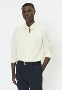 Chemise boutonnée jaune clair avec col à revers, manches retroussées, associée à un pantalon foncé à fines rayures et une ceinture noire tressée.
