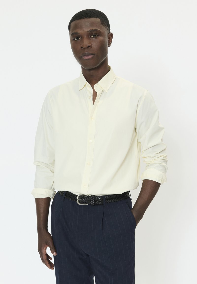 Chemise boutonnée jaune clair avec col à revers, manches retroussées, associée à un pantalon foncé à fines rayures et une ceinture noire tressée.