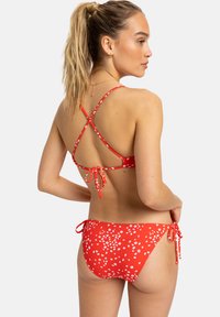 Red bikini with white floral pattern, featuring a crisscross back design, adjustable ties at the hips, and a smooth, stretchy fabric texture.