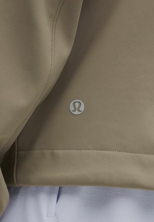 Beige Sportjacke mit glatter Textur, ausgestattet mit einem silbernen Logo auf der unteren Seite und einer sauberen Naht entlang dem Saum.