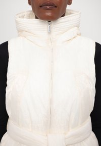 White padded vest with a high collar, zip front, and belted waist. Features a faint logo pattern and a smooth, shiny texture.