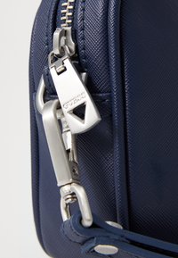 Borsa a tracolla in blu navy con texture, dotata di cerniera e accessori in argento. Presenta una tracolla robusta con chiusura a scatto e un piccolo tirazip a forma di triangolo.