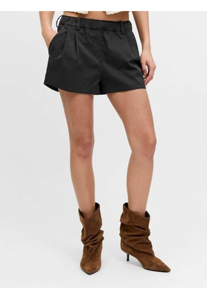 Vrouw draagt zwarte shorts met een hoge taille met zakken en bruine losse suède enkellaarsjes met lage hakken.